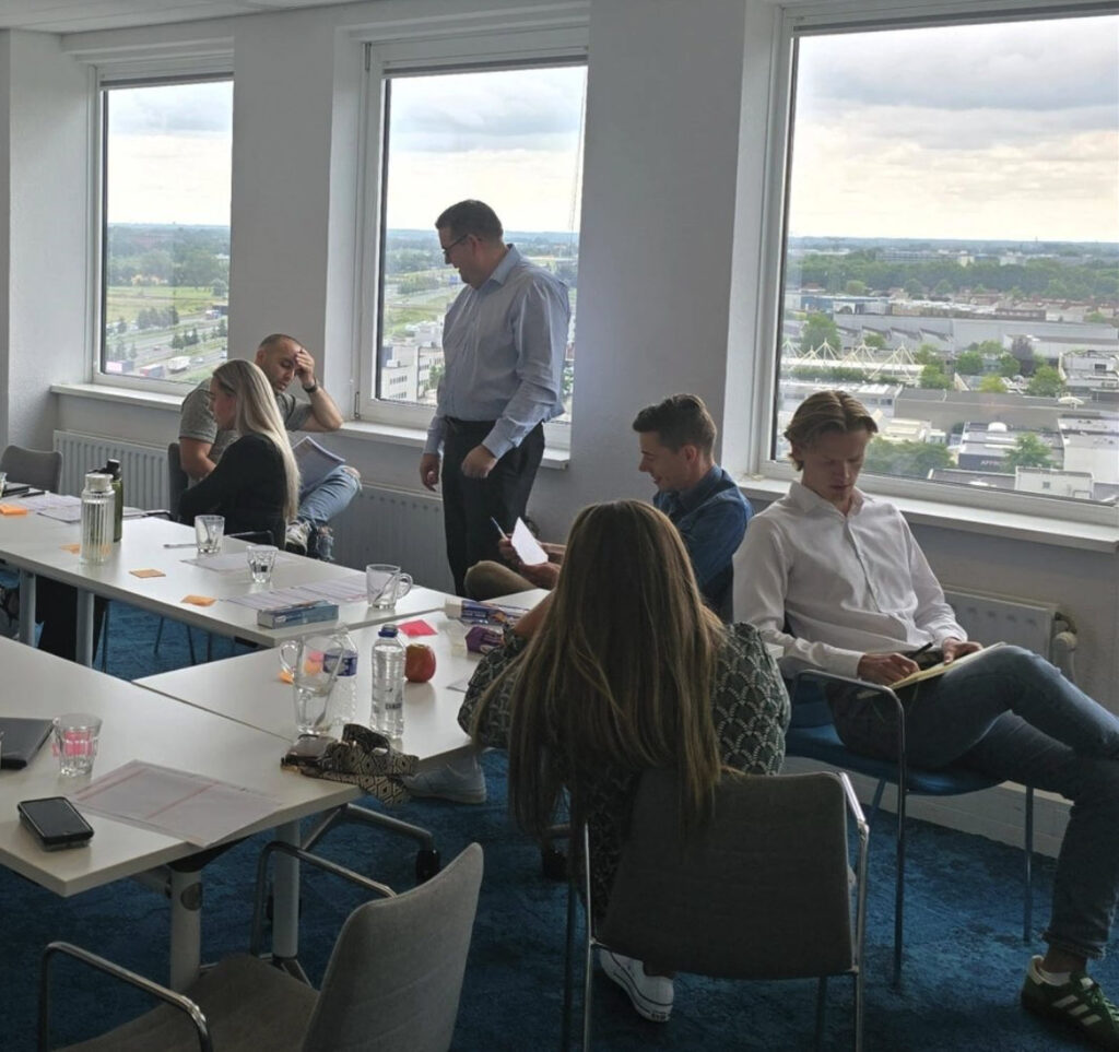 Andy McConville standing in a blue shirt and suit trousers, talking with an attendee in a training room, with big windows and a city view behind. (C) Andy McConville