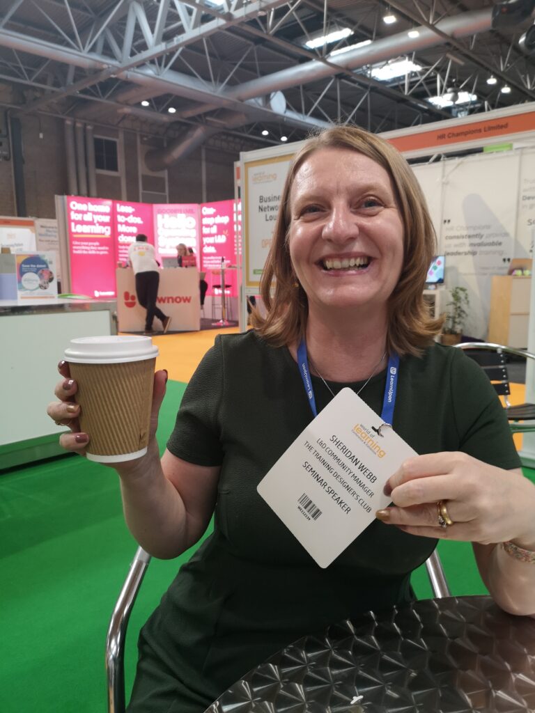 Sheridan Webb at the World of Learning conference, holding her name badge and a coffee, with stands in the background (C) Sheridan Webb