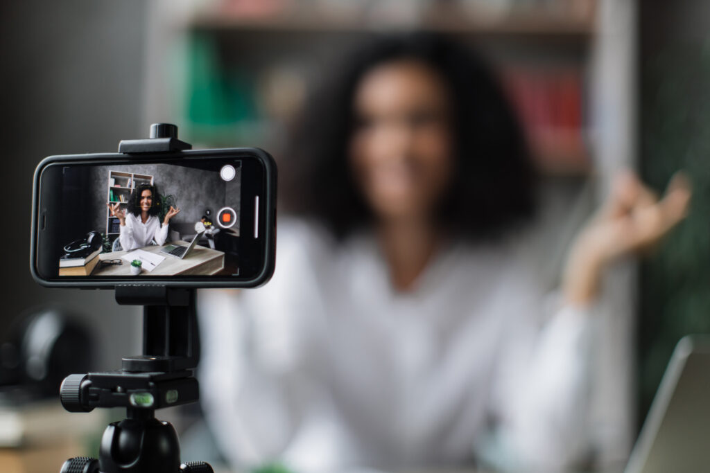 Blurred relaxed young woman in casual wear sitting on workplace with modern laptop and meditating with closed eyes during recording video blog. Focus on modern smartphone fixed on tripod.