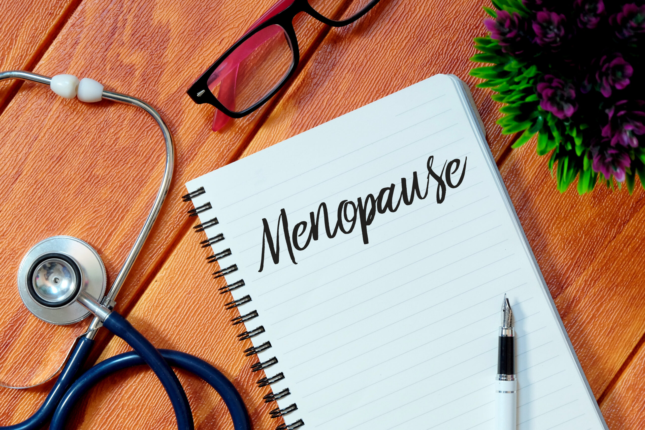 Top view of stethoscope,pink eyeglasses,plant,pen and notebook written with Menopause on wooden background.Health concept.