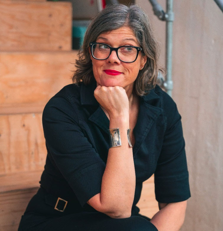 Kirsty Lewis - white woman with slightly grey chin length hair, wearing glasses and red lipstick, smiling, sitting on steps.