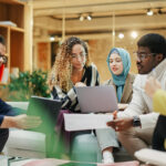 A Group of Multiethnic People Brainstorming Using Laptop and Charts in a Meeting Room at Office. Enthusiastic Young Adults Analyzing New Data To Improve Startup Performance. Medium Shot