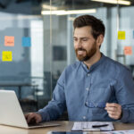 Smiling businessman working on laptop in modern office with colorful sticky notes on glass wall