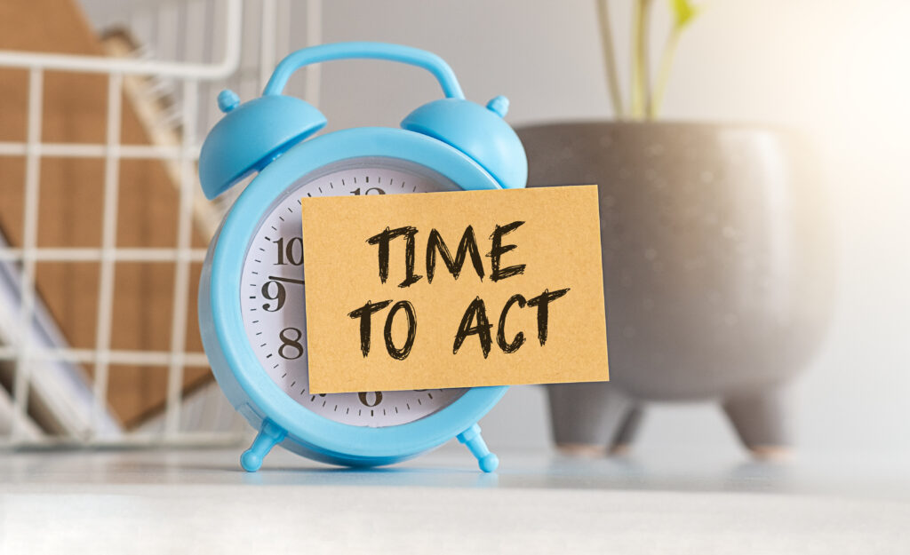 Blue alarm clock on desk with Time to Act sticky note and plant in background