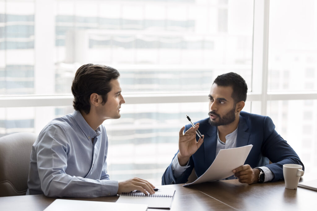 Two diverse serious young male business partners discussing contract terms, agreement text, marketing financial report, reviewing paper document at workplace table together